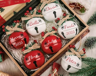 Puede incluir: Una caja de madera llena de adornos navideños. Los adornos tienen forma de campana, algunos rojos y otros blancos. Cada campana tiene un nombre y un diseño de copo de nieve. Algunas campanas dicen "Merry 2025". Las campanas están decoradas con lazos y bayas.