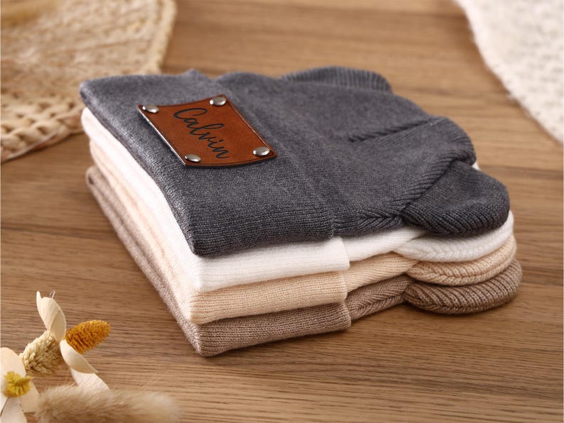 K&ouml;nnte beinhalten: Ein Stapel gefalteter M&uuml;tzen in verschiedenen neutralen Farben, darunter Grau, Wei&szlig; und Beige. Ein braunes Lederetikett mit dem Namen "Calvin" ist an der oberen grauen M&uuml;tze befestigt. Die M&uuml;tzen sind ordentlich auf einer Holzoberfl&auml;che angeordnet.