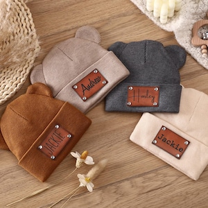 Puede incluir: Cuatro gorros de bebé personalizados en marrón, beige, gris y crema, cada uno con un parche de cuero que muestra un nombre. Los gorros tienen orejas de oso y están colocados sobre una superficie de madera con juguetes para bebés.