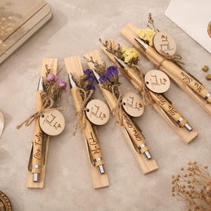 Könnte beinhalten: Fünf Bambuskugelschreiber mit silbernen Akzenten, die jeweils auf einem schmalen Holzsockel liegen. Jeder Stift ist mit einem kleinen Bündel Trockenblumen, einer Schnur und einem runden Holzanhänger mit der Gravur Thank you und Herz verziert.