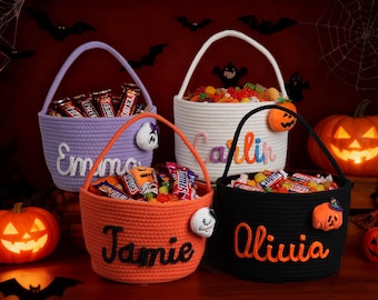 May include: Four Halloween-themed candy baskets in purple, orange, white, and black, each with a handle and personalized name embroidery. The baskets are filled with candy and decorated with Halloween-themed embellishments like pumpkins and ghosts.