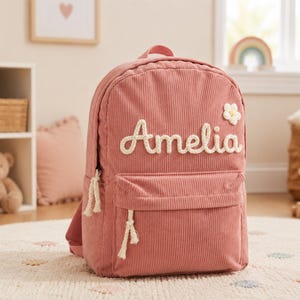 Könnte beinhalten: Ein altrosa Rucksack aus Kordstoff mit dem Namen Amelia, der in weißer, seilartiger Schrift auf der Vorderseite aufgestickt ist. Der Rucksack verfügt über eine kleine weiße Blumenapplikation, eine Fronttasche mit Reißverschluss und weiße Kordelanhänger.