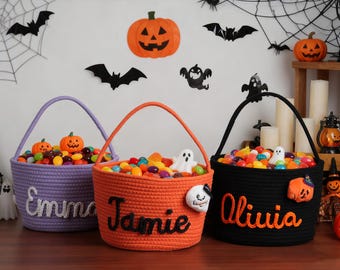 May include: Three Halloween-themed baskets in purple, orange, and black, filled with colorful candies. Each basket has a name embroidered on it: Emma, Jamie, and Olivia. Decorative pumpkins and ghosts are attached to the baskets.