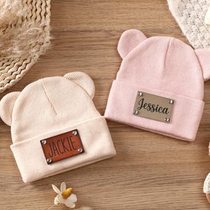 K&ouml;nnte beinhalten: Zwei Baby-M&uuml;tzen mit B&auml;renohren. Eine ist hellrosa, die andere beige. Jede hat ein braunes Kunstleder-Patch mit einem Namen: "Jessica" und "Jackie". Die H&uuml;te liegen auf einer Holzoberfl&auml;che.