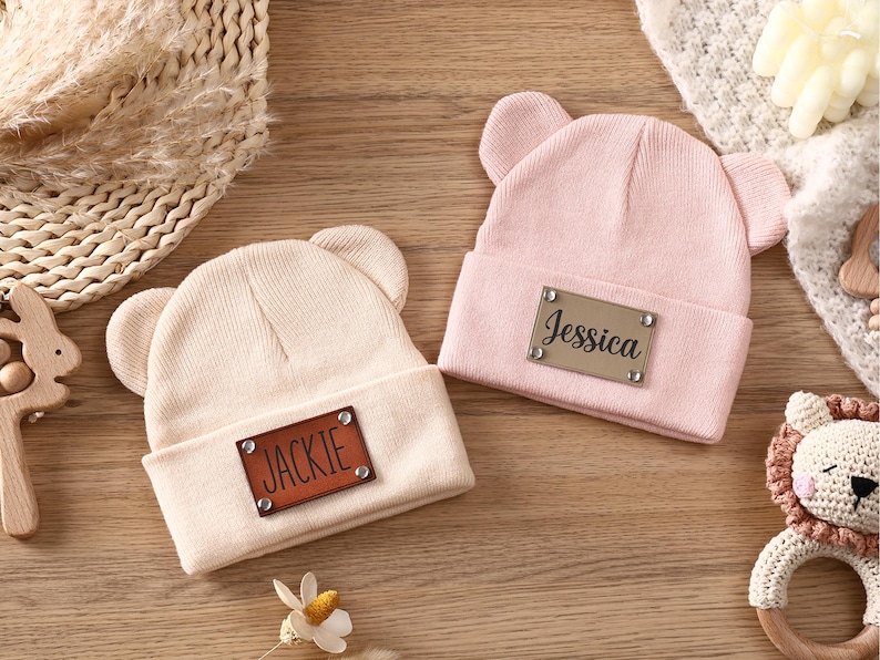 K&ouml;nnte beinhalten: Zwei Baby-M&uuml;tzen mit B&auml;renohren. Eine ist hellrosa, die andere beige. Jede hat ein braunes Kunstleder-Patch mit einem Namen: "Jessica" und "Jackie". Die H&uuml;te liegen auf einer Holzoberfl&auml;che.