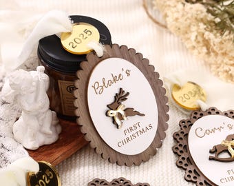 May include: Wooden Christmas ornament with an oval shape and a scalloped edge. The ornament features the name "Blake's", a reindeer, and the text "FIRST CHRISTMAS". A gold-colored tag with the year "2025" is attached with a white ribbon.