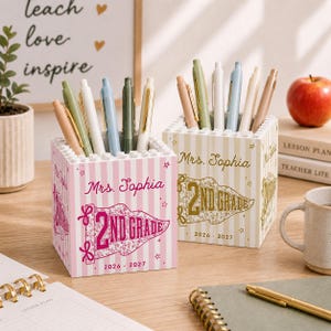 Puede incluir: Dos portalápices cuadrados con un borde superior tipo bloque. Uno es de rayas rosas y blancas con el texto "Mrs. Sophia" y "2ND GRADE 2026 - 2027" en rosa. El otro es de rayas beige y blancas con el mismo texto en dorado. Ambos contienen bolígrafos.