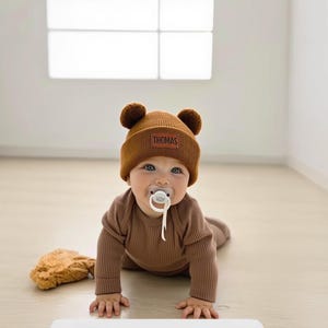 K&ouml;nnte beinhalten: Ein Baby mit einem braunen B&auml;renhut mit dem Namen "THOMAS", einem passenden braunen Strampler und einem Schnuller. Das Baby krabbelt auf einem hellen Boden. Ein Stofftier liegt neben dem Baby.