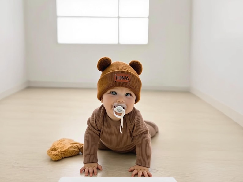 K&ouml;nnte beinhalten: Ein Baby mit einem braunen B&auml;renhut mit dem Namen "THOMAS", einem passenden braunen Strampler und einem Schnuller. Das Baby krabbelt auf einem hellen Boden. Ein Stofftier liegt neben dem Baby.