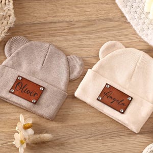 K&ouml;nnte beinhalten: Zwei beige gestrickte Baby-M&uuml;tzen mit B&auml;renohren. Jede M&uuml;tze hat ein braunes Lederpatch mit einem Namen: "Oliver" und "Angela". Die M&uuml;tzen liegen auf einer Holzoberfl&auml;che.