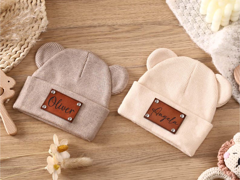 K&ouml;nnte beinhalten: Zwei beige gestrickte Baby-M&uuml;tzen mit B&auml;renohren. Jede M&uuml;tze hat ein braunes Lederpatch mit einem Namen: "Oliver" und "Angela". Die M&uuml;tzen liegen auf einer Holzoberfl&auml;che.