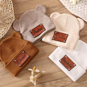 K&ouml;nnte beinhalten: Vier gestrickte Baby-Beanies in Beige, Braun, Hellbraun und Wei&szlig;, jeweils mit einem Lederaufn&auml;her. Die Aufn&auml;her sind mit den Namen "Andrew", "Barton", "Daniel" und "Jackie" personalisiert. Die Beanies haben B&auml;renohren.