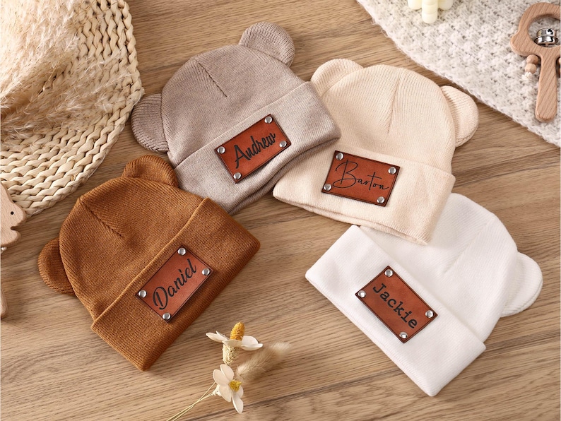 K&ouml;nnte beinhalten: Vier gestrickte Baby-Beanies in Beige, Braun, Hellbraun und Wei&szlig;, jeweils mit einem Lederaufn&auml;her. Die Aufn&auml;her sind mit den Namen "Andrew", "Barton", "Daniel" und "Jackie" personalisiert. Die Beanies haben B&auml;renohren.