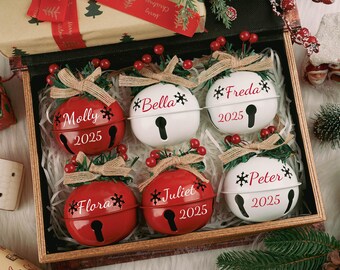 Puede incluir: Una caja de madera contiene seis adornos de campana navideños personalizados. Tres son rojos, tres son blancos, cada uno con un nombre, un copo de nieve y el año 2025. Están adornados con lazos de arpillera y bayas artificiales, ideales para la decoración navideña.