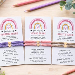 May include: Three white rectangular cards featuring a rainbow graphic and text. Each card holds two woven bracelets with a small gold heart charm. The cards are labeled First Day of Kindergarten, Second Grade, and Back to School with poems.