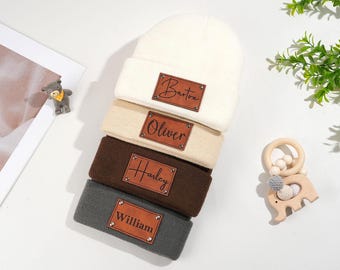 Pode incluir: Uma pilha de quatro gorros personalizados em creme, bege, castanho e cinzento. Cada gorro tem um remendo de couro castanho com um nome em escrita: Barton, Oliver, Harley e William. Um brinquedo de madeira para bebé e uma pequena figura de urso também estão na imagem.