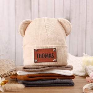 K&ouml;nnte beinhalten: Ein Stapel Baby-M&uuml;tzen in verschiedenen Farben, darunter Beige, Braun, Wei&szlig;, Orange und Grau. Die oberste M&uuml;tze hat B&auml;renohren und ein braunes Lederetikett mit der Aufschrift "THOMAS". Die M&uuml;tzen sind aus weichem Strickmaterial.
