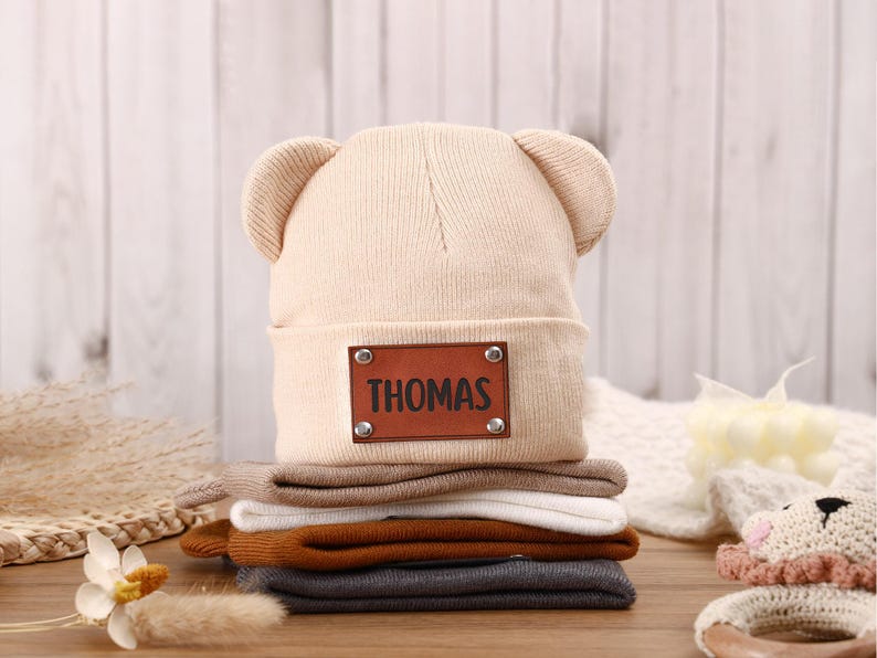 K&ouml;nnte beinhalten: Ein Stapel Baby-M&uuml;tzen in verschiedenen Farben, darunter Beige, Braun, Wei&szlig;, Orange und Grau. Die oberste M&uuml;tze hat B&auml;renohren und ein braunes Lederetikett mit der Aufschrift "THOMAS". Die M&uuml;tzen sind aus weichem Strickmaterial.
