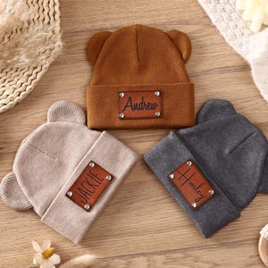 K&ouml;nnte beinhalten: Drei personalisierte Baby-Beanies in Braun, Beige und Grau, jeweils mit B&auml;renohren. Die braune M&uuml;tze hat "Andrew" auf einem Lederpatch, die beige "Jackie" und die graue "Harley". Sie werden auf einer Holzoberfl&auml;che pr&auml;sentiert.