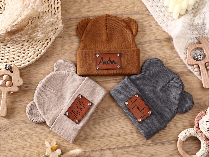 K&ouml;nnte beinhalten: Drei personalisierte Baby-Beanies in Braun, Beige und Grau, jeweils mit B&auml;renohren. Die braune M&uuml;tze hat "Andrew" auf einem Lederpatch, die beige "Jackie" und die graue "Harley". Sie werden auf einer Holzoberfl&auml;che pr&auml;sentiert.