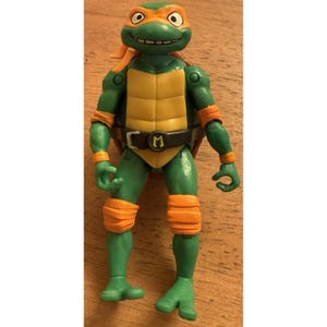 May include: A green action figure of a Teenage Mutant Ninja Turtle, featuring orange accents on the mask, knee pads, and arm bands. The figure has a yellow plastron and a black belt with a letter "M".