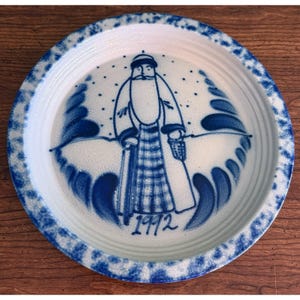 May include: A round ceramic plate with a white base and a blue sponge-painted rim. The centre features a blue hand-painted illustration of a bearded figure in a long coat and hat, surrounded by dots and stylised waves, with the year 1992 written below.