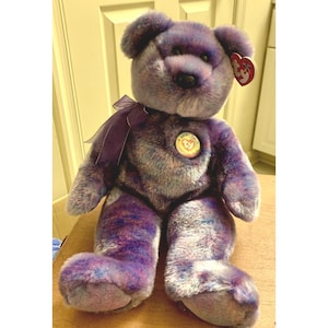 May include: A plush purple teddy bear with a ribbon around its neck and a heart-shaped tag. The bear has a round, button nose and a circular emblem on its chest. The fur is a mix of purple, blue, and gray.