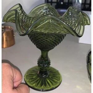 Könnte beinhalten: Grüne Glas-Fußschale mit gewelltem Rand. Die Schale hat ein Rautenmuster am Körper und einen runden Sockel mit Sternmuster. Der Stiel hat ein dekoratives, bauchiges Element.