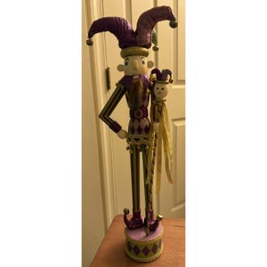 May include: A decorative wooden jester nutcracker figurine. The jester wears a purple hat with bells, a green and purple striped outfit, and a matching base. The figurine holds a smaller jester doll.
