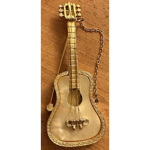 May include: A gold-toned guitar-shaped brooch with a pearlescent face. The brooch features a detailed design, including strings, a bridge, and a decorative border. A gold chain is attached to the top of the brooch. The brooch is likely made of metal.