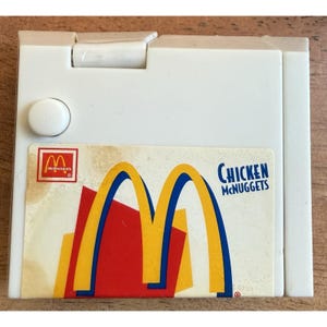 May include: A white plastic McDonald's Chicken McNuggets toy. The toy has a hinged lid and a button on the side. The front features the McDonald's logo and the words "Chicken McNuggets". The logo has yellow arches, red and yellow shapes.