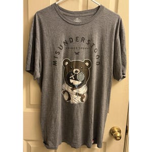 May include: A gray t-shirt with a graphic of a bear wrapped in bandages. The bear has a spiral eye and a cross on its face. The text "MISUNDERSTOOD" and "SINCE 1999" are printed on the shirt.