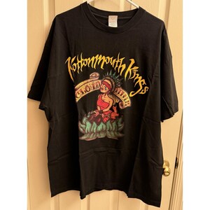 May include: Black t-shirt featuring the text "Kottonmouth Kings" in yellow and a graphic of a figure in red and white on a lotus flower. The shirt is hanging on a white hanger.