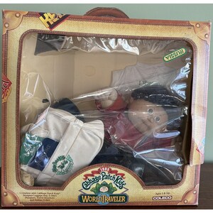 May include: A 1985 Cabbage Patch Kids World Traveler doll set in a suitcase-style box. The set includes a doll with dark hair wearing a red shirt, a white canvas travel bag with a green logo, a passport, a travel brochure, and an airline ticket.