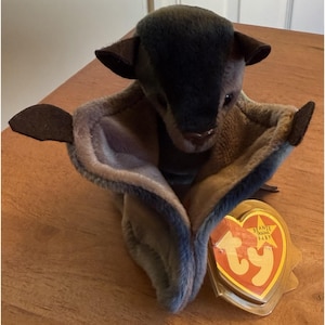 May include: A plush toy bat with a dark gray head and brown ears and tail. The bat's wings are a mix of gray and tan. A yellow and red TY Beanie Baby tag is attached to the toy.