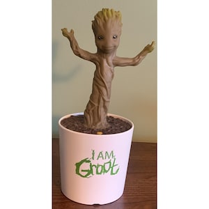 May include: A brown, tree-like figure with a textured bark surface and raised arms stands in a white cylindrical pot. The pot features the green text I AM GROOT on the front. The base of the figure is surrounded by dark brown, textured soil.