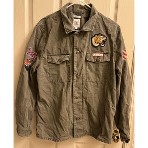 Könnte beinhalten: Olivgrüne Knopfjacke mit Kragen, zwei Brusttaschen und langen Ärmeln. Die Jacke hat Aufnäher, darunter einen Tiger, einen Sonnenuntergang und eine amerikanische Flagge. Der Markenname "GAP" ist auf dem Kragen sichtbar.