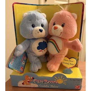 May include: Two plush Care Bears, a light blue bear with a rain cloud belly and a pink bear with a rainbow belly, are displayed in a box. The box reads "Care Bears Cuddle Pairs" and "Cheer Bear Grumpy Bear."