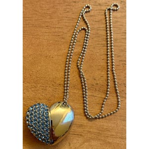 May include: A silver-toned heart-shaped pendant necklace featuring a ball chain. One half of the heart is smooth and reflective, while the other half is encrusted with numerous small, light blue circular crystals arranged in a grid-like pattern.