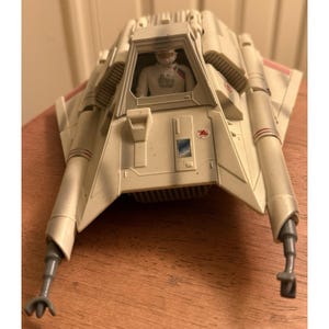 May include: A vintage toy model of a snowspeeder, primarily off-white with red accents. The cockpit features a pilot figure. The vehicle has two front prongs and two side wings. The toy is made of plastic.