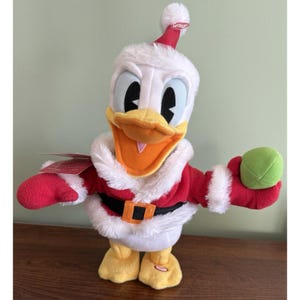 May include: A plush Donald Duck dressed in a Santa Claus outfit. The duck is wearing a red and white Santa suit with a black belt and a matching hat. It is holding a green ball in one hand.