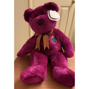 Puede incluir: Oso de peluche morado con una cinta marrón atada alrededor del cuello. El oso tiene una etiqueta adherida a su oreja y un pequeño parche bordado con un diseño de globo. El oso está sentado sobre una superficie de madera.