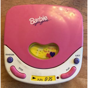 May include: A pink and white portable CD player with the Barbie for girls logo printed on the top lid. The device features a heart-shaped viewing window, a digital LCD screen displaying play time, and purple and pink control buttons for functions.