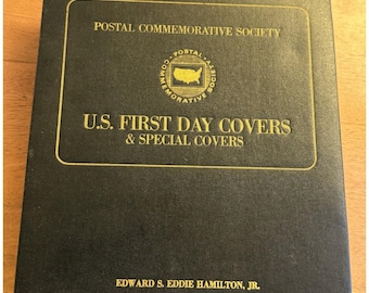 May include: A black album with gold lettering and a gold-framed emblem. The text reads "POSTAL COMMEMORATIVE SOCIETY" and "U.S. FIRST DAY COVERS & SPECIAL COVERS." The album is for collecting stamps and covers.