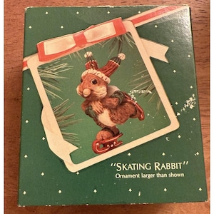 Peut inclure: Ornement de Noël représentant un lapin brun portant un chapeau et des patins rouges et blancs. Le lapin est sur une boîte verte avec un cadre blanc et un nœud rouge. Le texte sur la boîte indique "SKATING RABBIT" et "Ornament larger than shown."