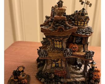 May include: A detailed Halloween-themed miniature house with a bat weathervane, carved pumpkins, and gargoyle statues. The house has multiple levels, a chimney, and a sign that reads "Come out in all bewitchery". A small figurine is also present.