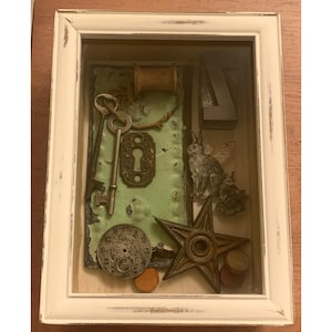 Könnte beinhalten: Ein gerahmter Schattenkasten mit einem weißen, abgenutzten Rahmen. Im Inneren sind verschiedene Vintage-Objekte auf einem grünen Hintergrund angeordnet, darunter Schlüssel, ein Schlüsselloch, ein Zifferblatt, ein Stern, eine Holzspule und eine geflügelte Kaninchenfigur.