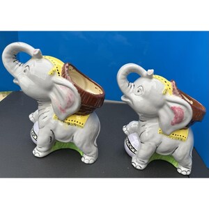 May include: Two grey ceramic elephant ornaments with brown baskets on their backs. The elephants have pink ears, yellow saddle blankets, and white tusks. The ornaments sit on a green base.