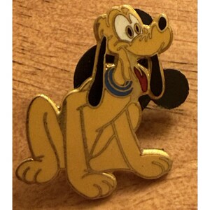 Puede incluir: Un pin de Pluto amarillo con orejas negras y una forma de cabeza de Mickey Mouse negra detrás. El pin presenta a Pluto sentado con un collar azul y una lengua roja. El pin está sobre una superficie de madera.