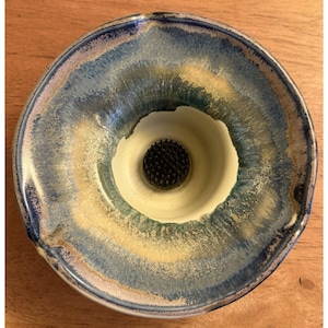 May include: A round ceramic flower frog bowl featuring a reactive glaze with blended shades of deep blue, teal, and cream. The center of the bowl contains a circular metal kenzan with numerous sharp, upright needles for arranging floral stems.